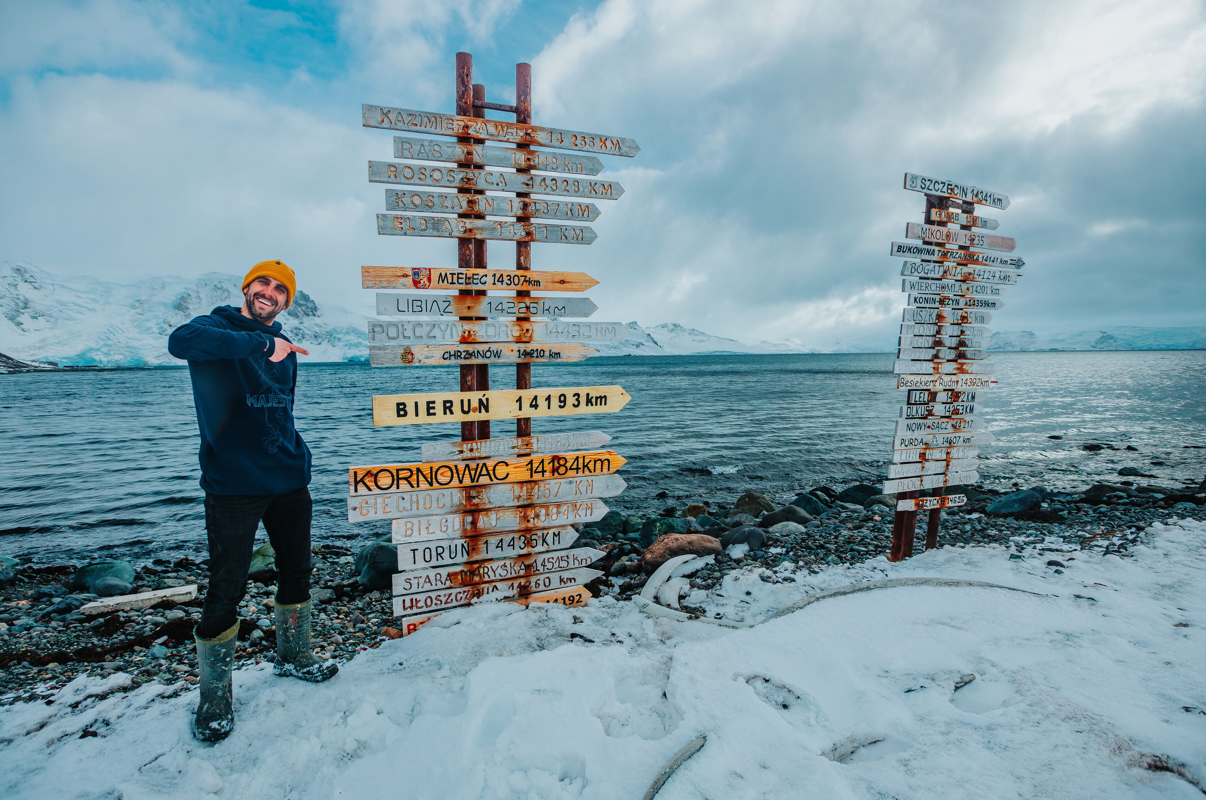 Młody, uśmiechnięty człowiek w pomarańczowej czapce stoi w śniegu na brzegu oceanu i pokazuje drogowskaz z nazwami polskich miast. W środku tabliczka: Bieruń 14193 km. W tle lodowe góry.