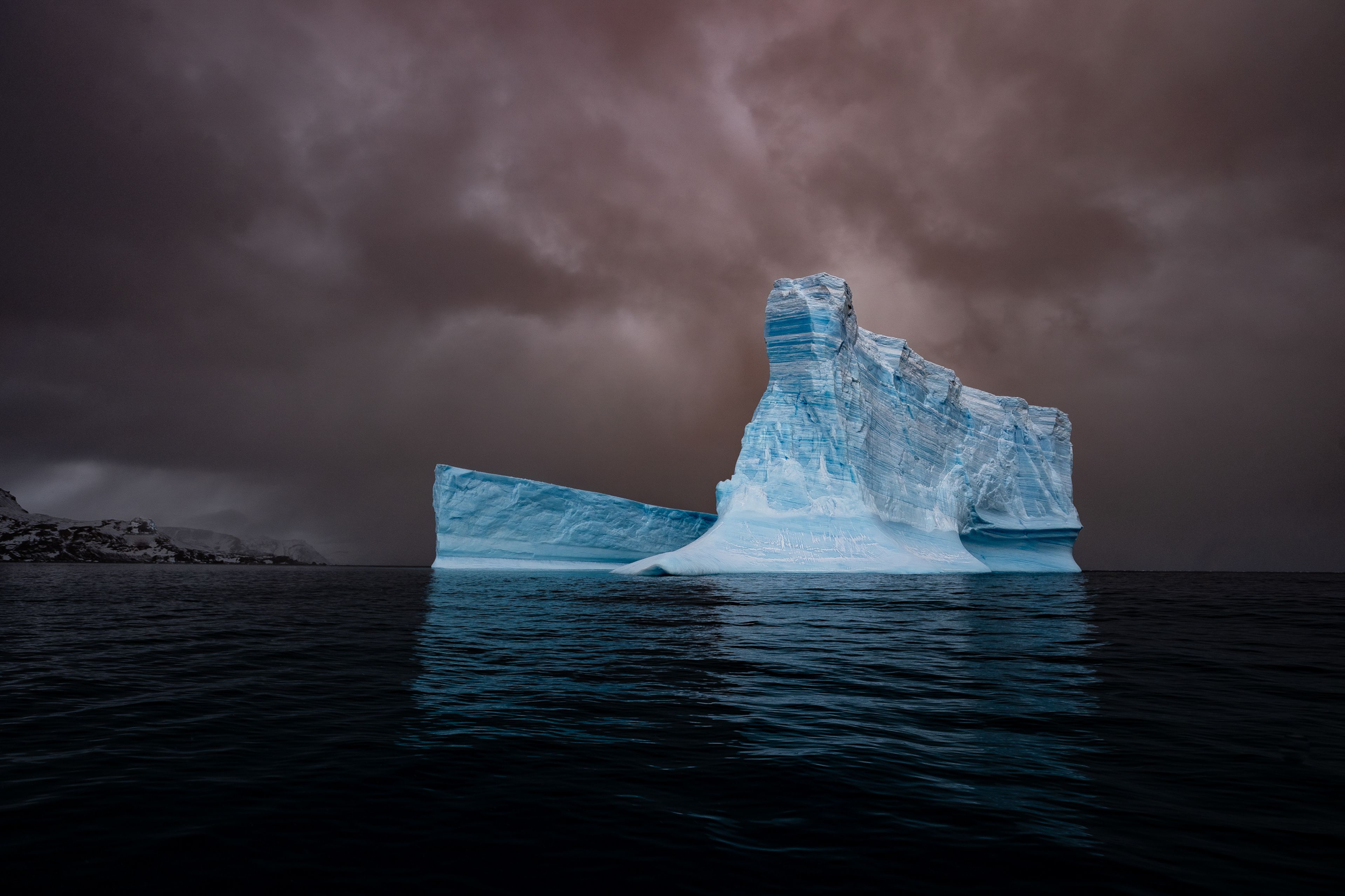 Niebieska góra lodowa dryfująca w oceanie na tle brunatnego nieba.