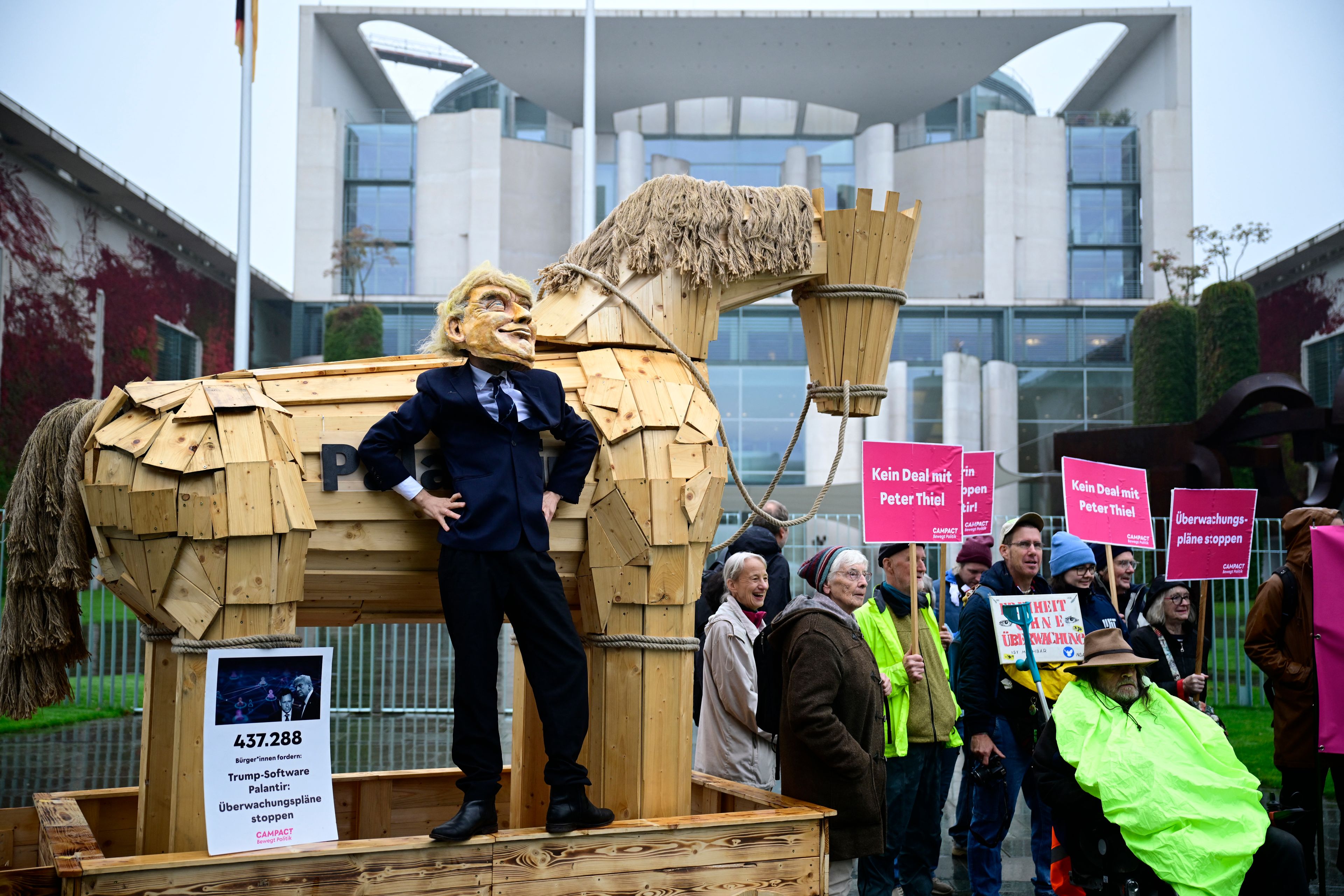 An activist of the group Campact! dressed as US President Donald Trump (L) stands next to a wooden horse symbolising a Trojan Horse with the lettering "Palantir" in front of the Chancellery in Berlin, where is taking place the German cabinet's weekly meeting on October 8, 2025. Activists protested against the German Interior Ministry's intention to pave the way for the nationwide use of the data mining Palantir software, that was developed by a company co-founded and chaired by Trump supporter Peter Thiel. (Photo by Tobias SCHWARZ / AFP)