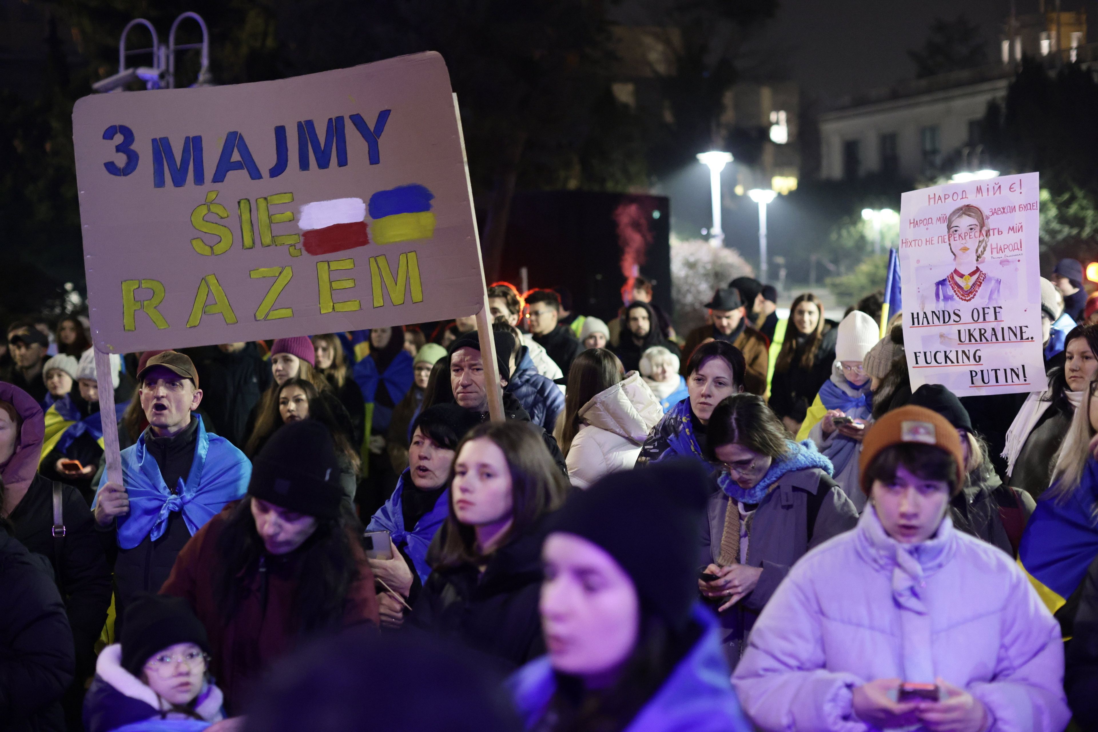 Na zdjęciu jest tłum aktywistów. Dwóch mężczyzn trzyma plakat z napisaem "Trzymajmy się razem". Słowa te są napisane w niebiesko-żółtych kolorach. Po prawej stronie narysowane są dwie flagi: biało-czerwona i niebiesko-żółta