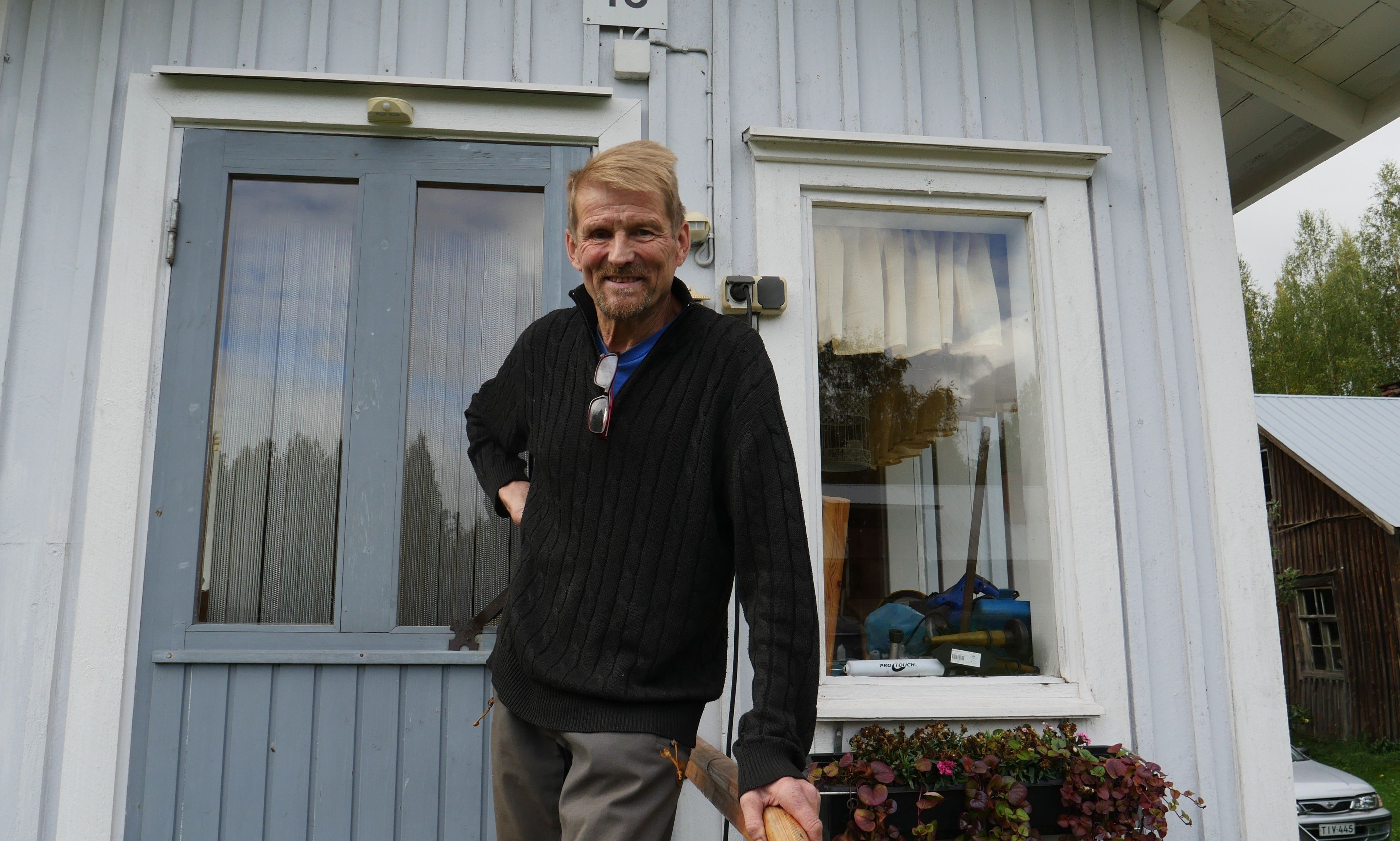 Zdjęcie mężczyzny o jasnych włosach ubranego w czarny sweter. Stoi przed domem oparty o poręcz.