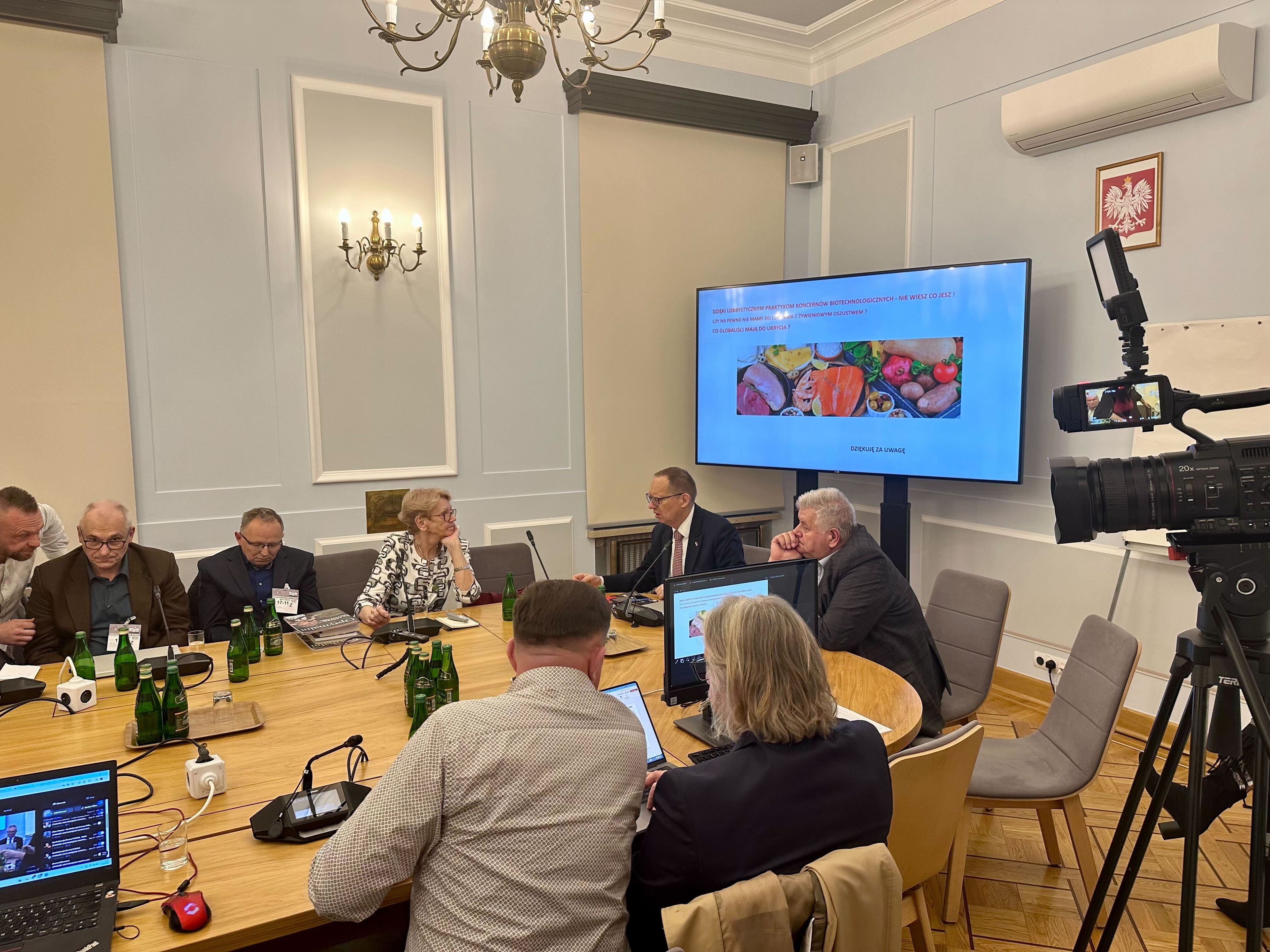 Grupa ludzi siedzących przy stole, oglądających prezentację na ekranie.