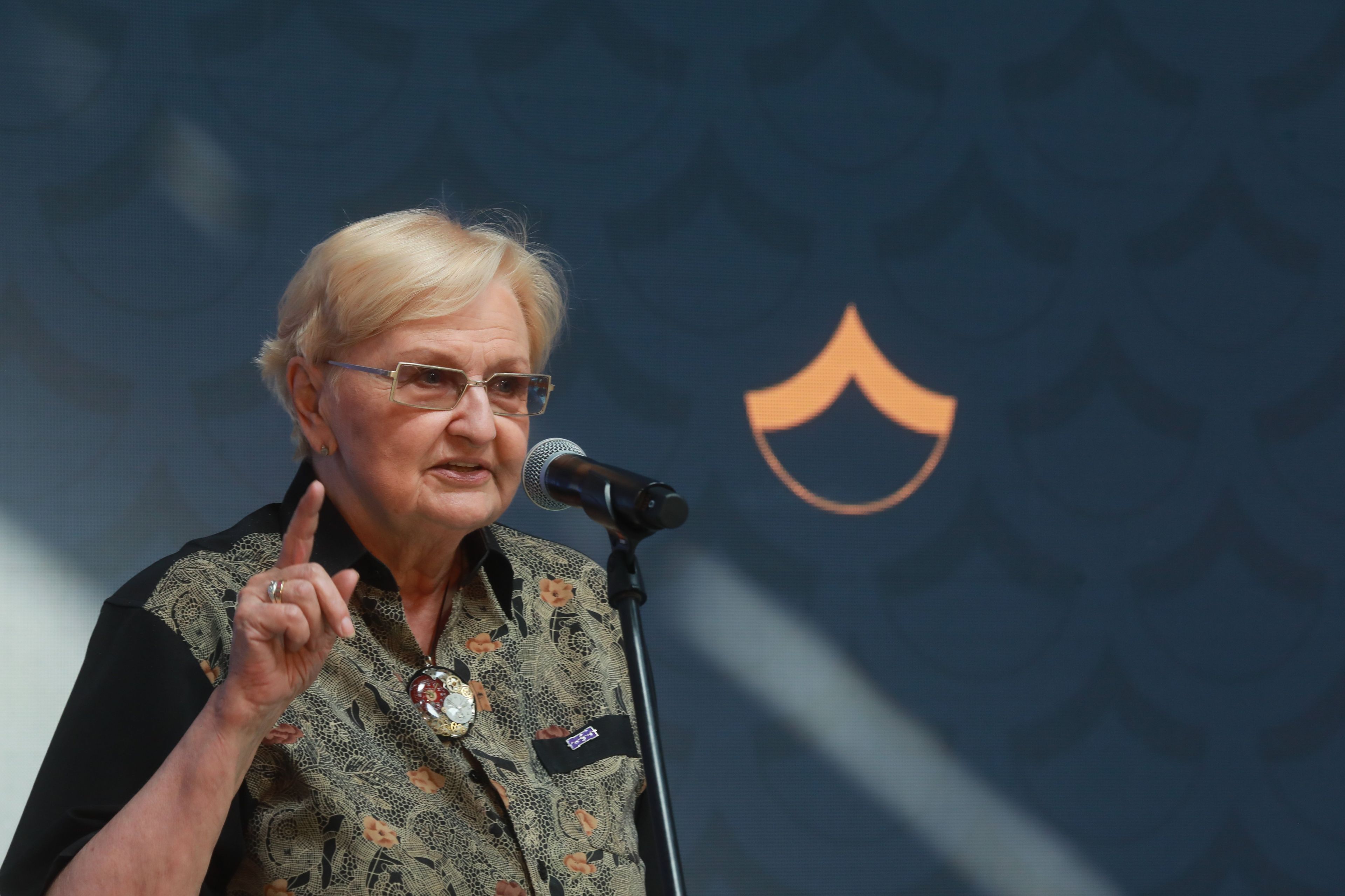 Kobieta we wzorzystej bluzce przemawia i unosi do góry wskazujący palec prawej ręki