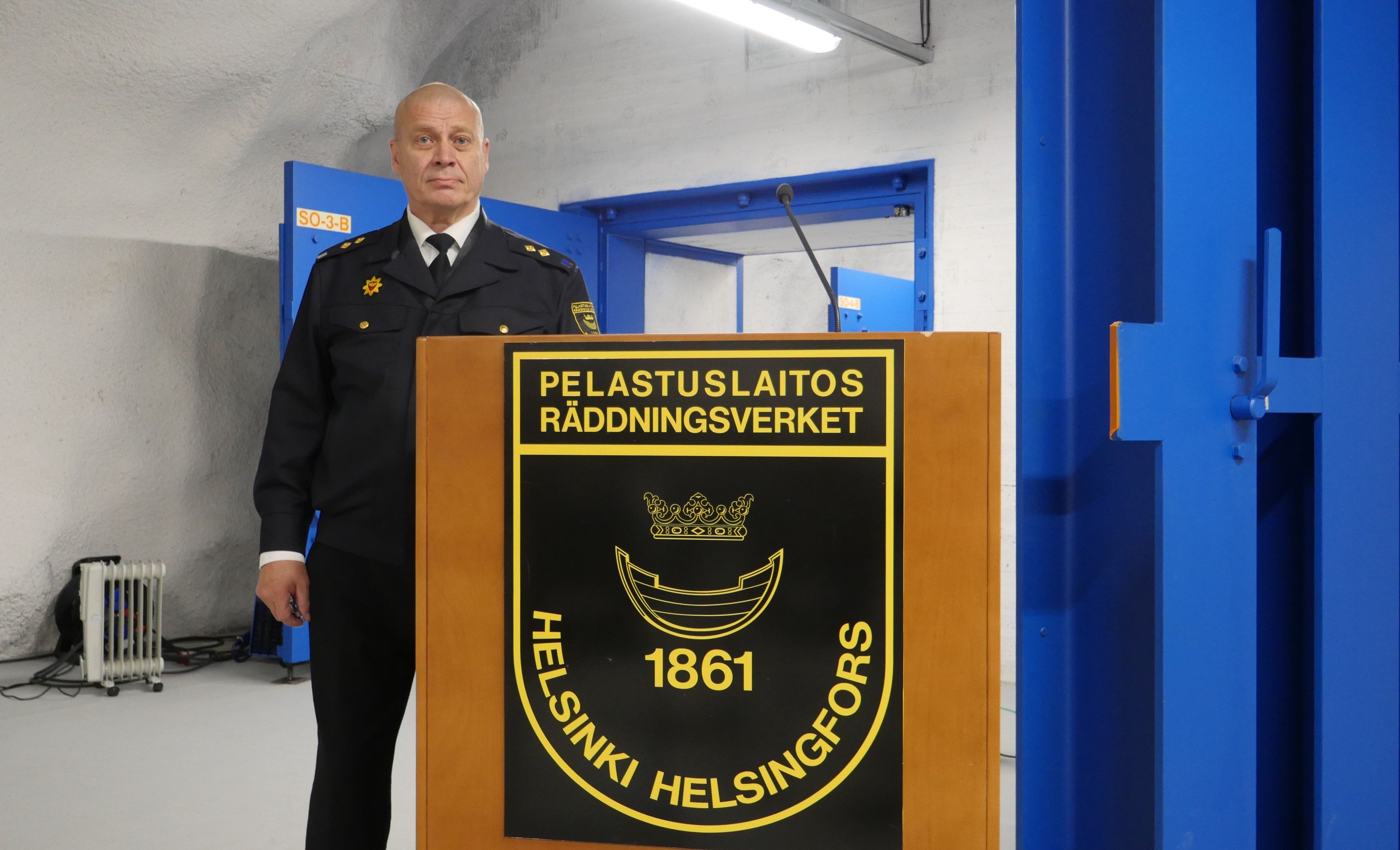 Mężczyzna w mundurze stoi w surowym pomieszczeniu z niebieskimi, masywnymi drzwiami. Pokazuje tablicę z napisami po fińsku i datą 1861 rok.
