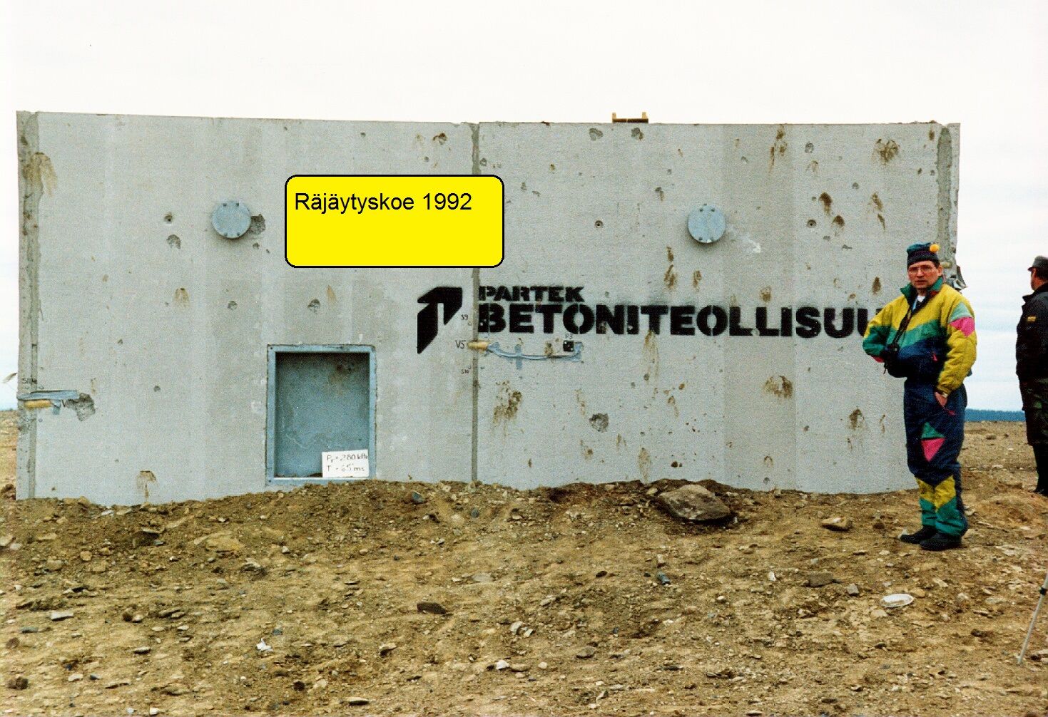 Mężczyzna w kolorowym kombinezonie stoi na tle betonowego budynku, dookoła rozkopana ziemia. Na fotografii data 1992