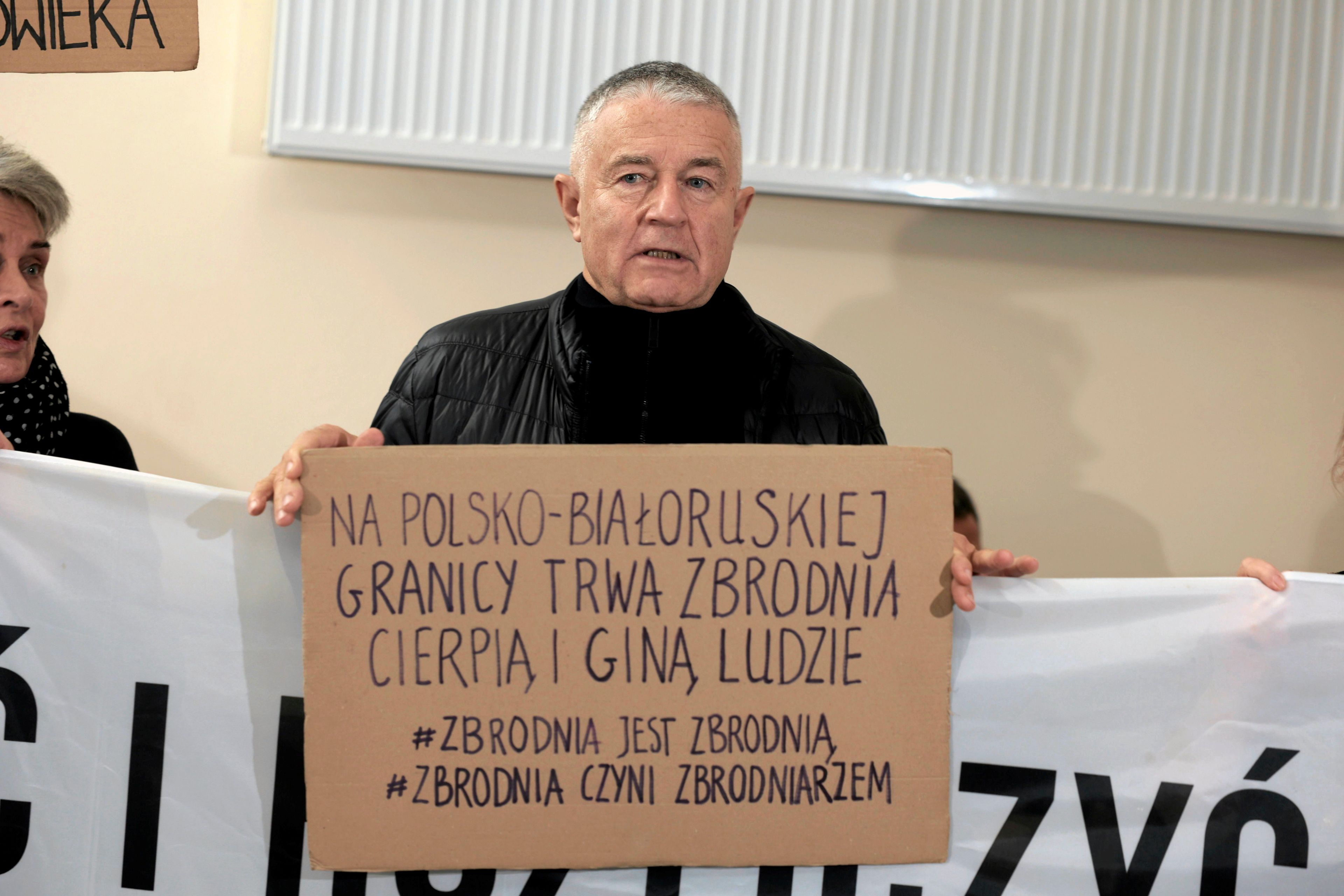 Siwy mężczyzna w czarnej kurtce (Władysław Frasyniuk) trzyma kawałek kartonu z napisem "Na polsko-białoruskiej granicy trwa zbrodnia. Cierpią i giną ludzie. #Zbrodnia jest zbrodnią #Zbrodnia czyni zbrodniarzem"