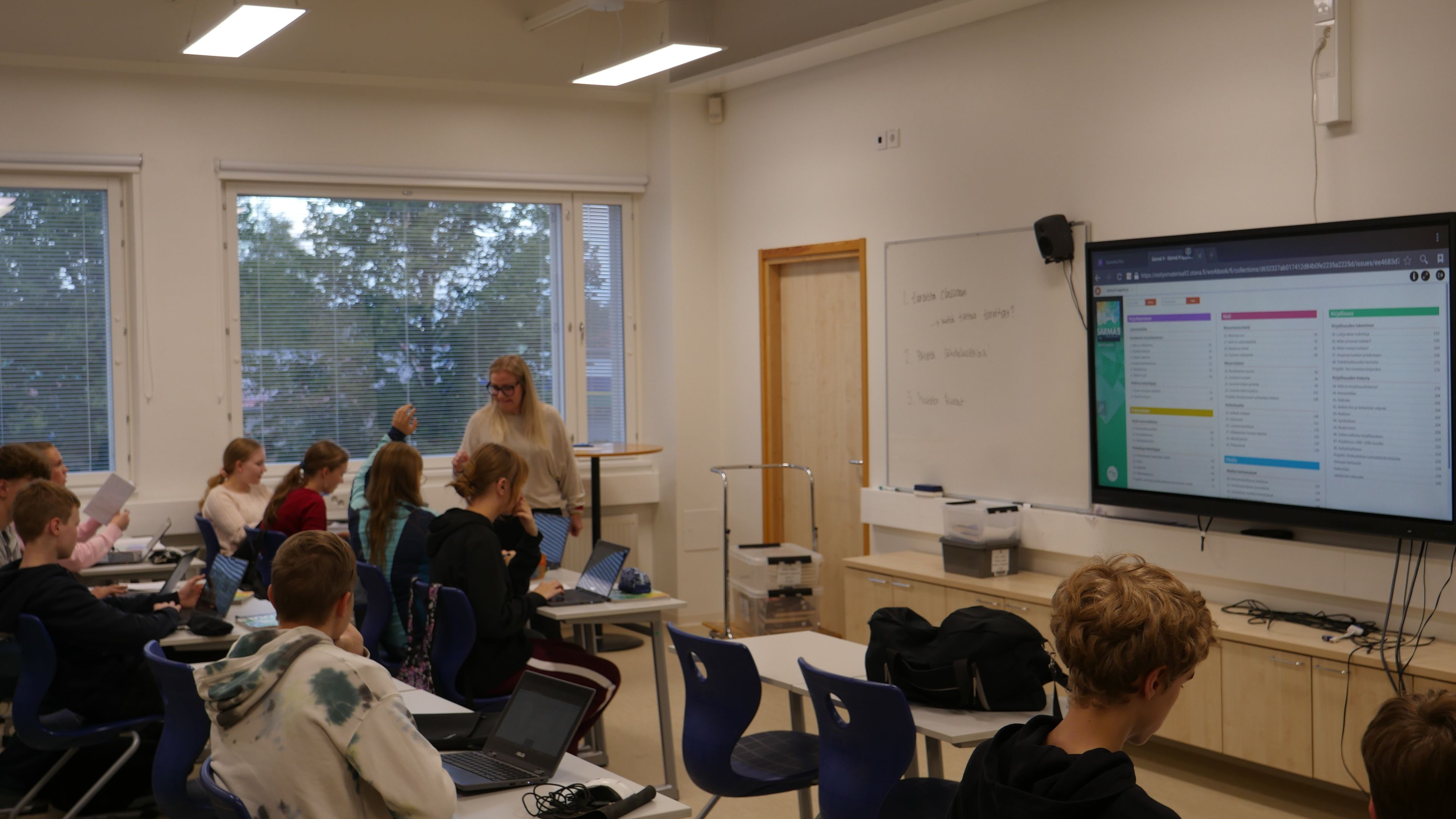 Szkolna klasa, lekcja, uczniowie z otwartymi laptopami śledzą ekran multimedialnej tablicy.