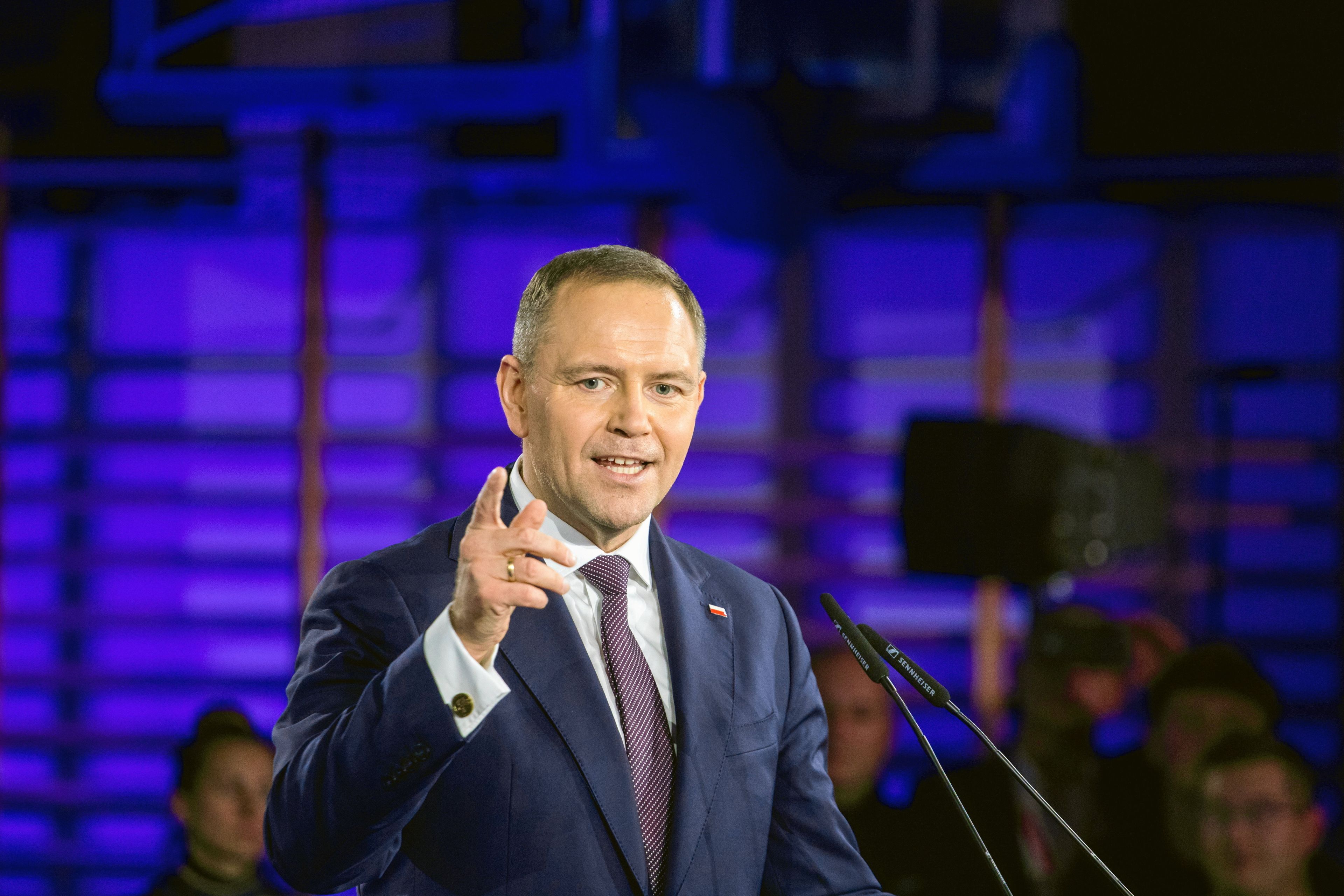 Mężczyzna w garniturze przemawia, wskazuje na cos ręką