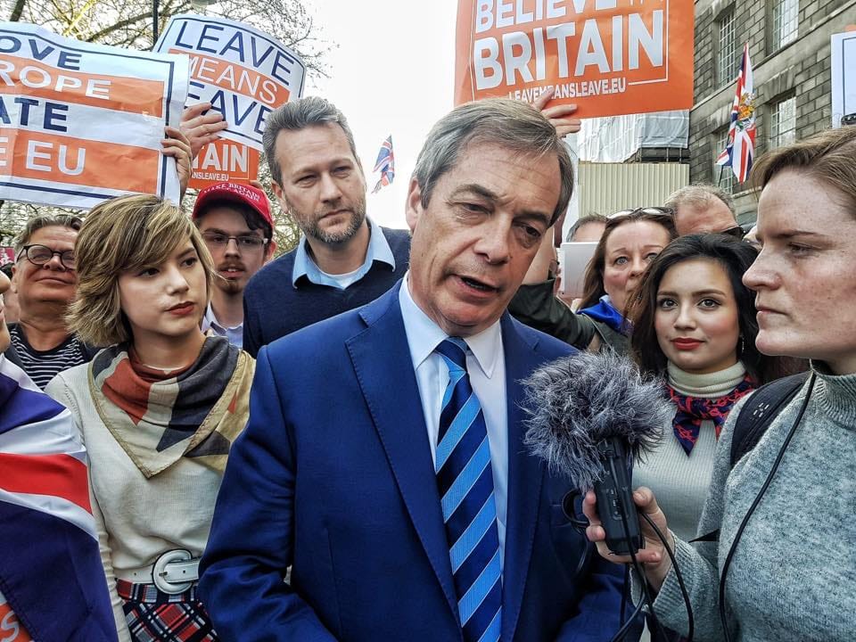 Mężczyzna w średnim wieku na tle osób z banerami namawiającymi do wyjścia Wielkiej Brytanii z UE. Za nim stoi Nathan Gill.