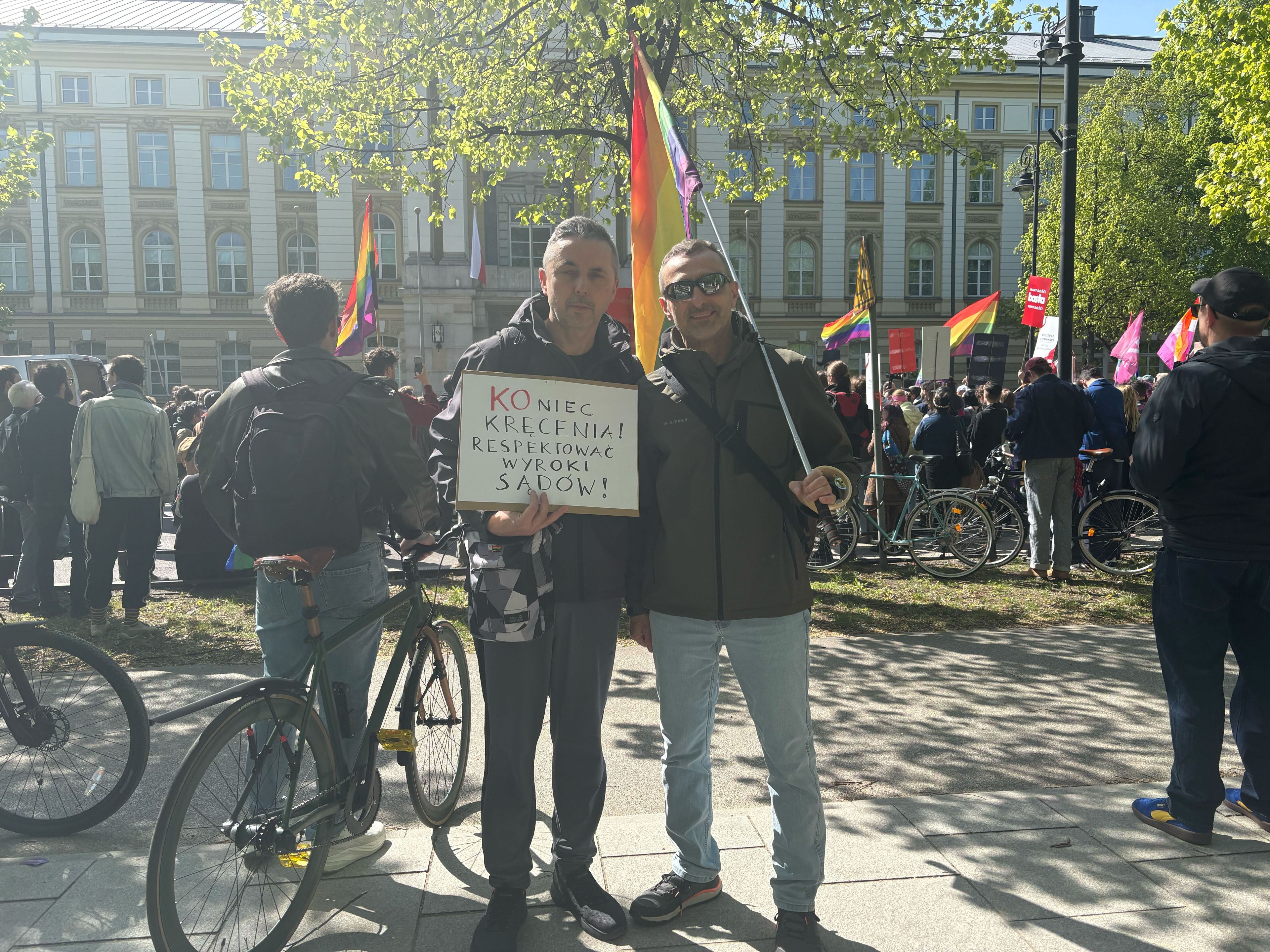 dwóch mężczyzn stojących razem, jeden trzyma tęczową flagę, drugi trzyma karton z napisem "koniec kręcenia! respektować wyroki sądów"