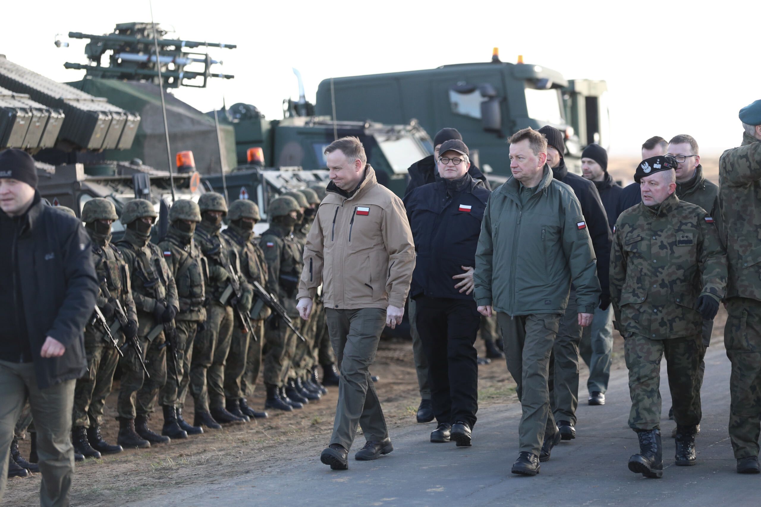 Zołnierze i urzednicy w wojskowych kurtkach na poligonie. W tle - broń