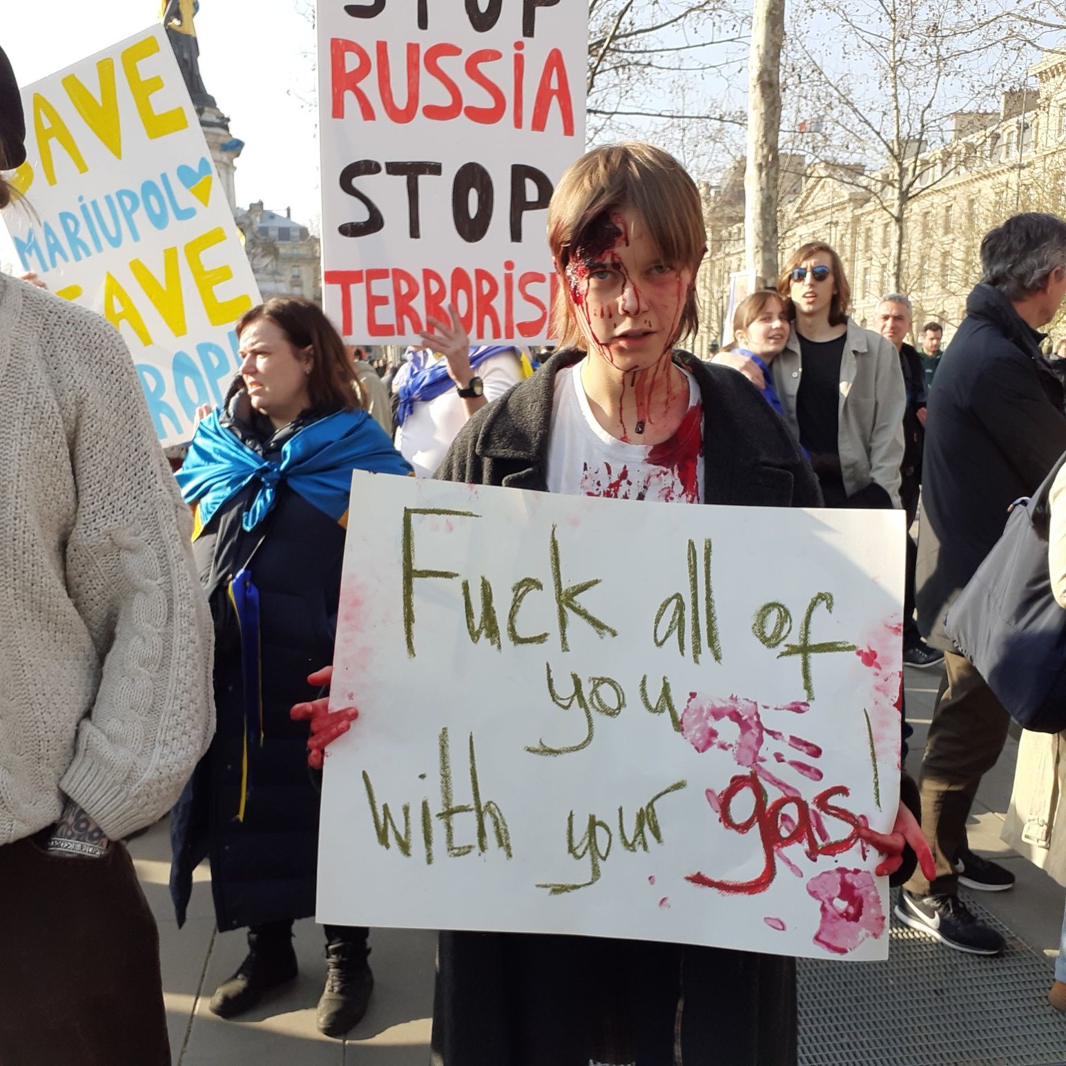 Chłopak z namalowanymi na twarzy śladami krwi trzyma baner z napisem "Fuck all of you with your gas"