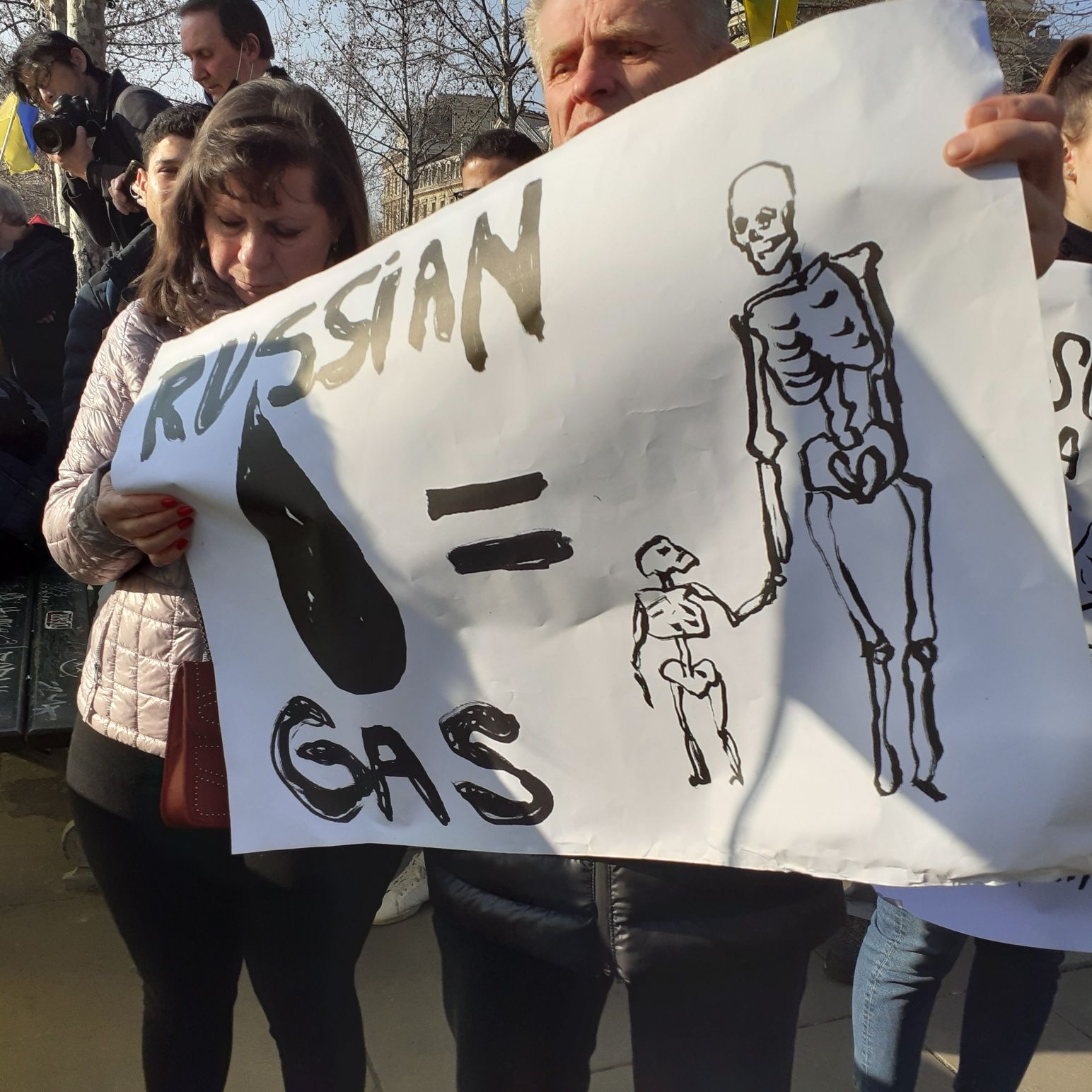 Demonstrant trzyma baner z napisem "Russian gas równa się sylwetka osoby dorosłej i dwójki dzieci, w domyśle uchodźcy