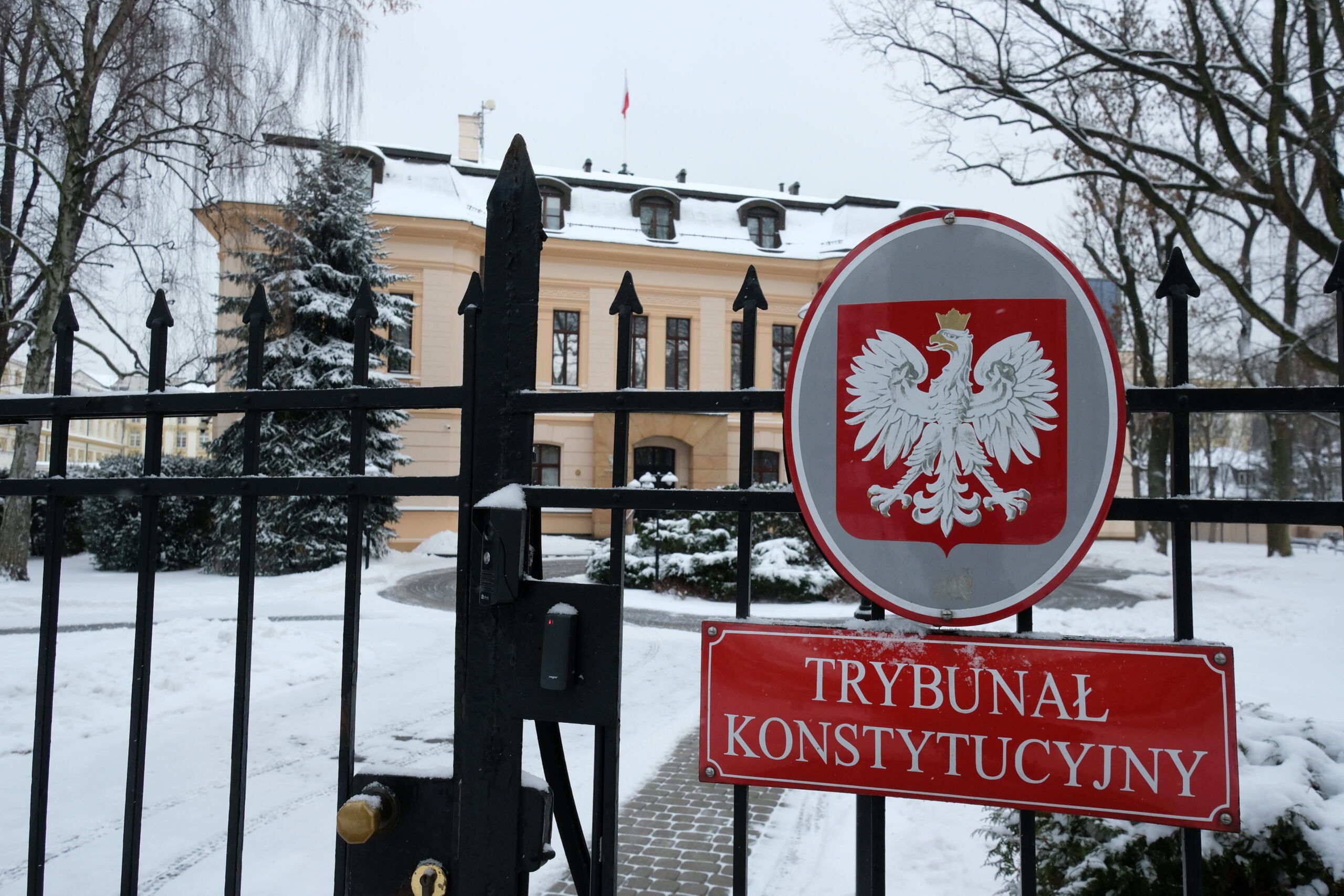 Tabliczka z napisem "Trybunał Konstytucyjny" na czerwonym tle. Na drugim planie budynek Trybunału
