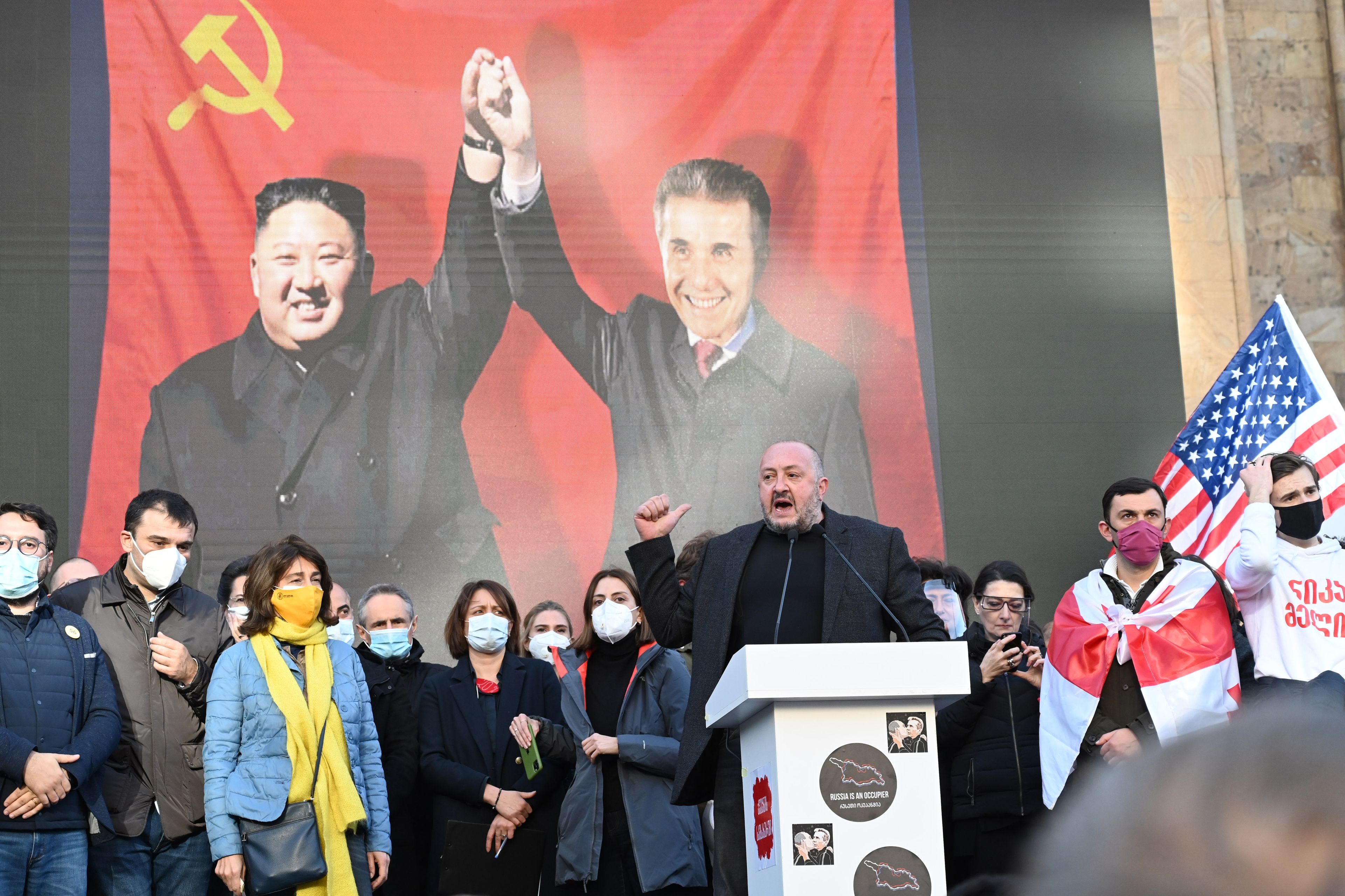 Ludzie w czasie protestu. Kobieta ubrana w barwy ukraińskie. W tle - plakat z gruzińskim politykiem trzymającym się za ręce z przywódcą Korei Północnej na tle sowieckiej flagi