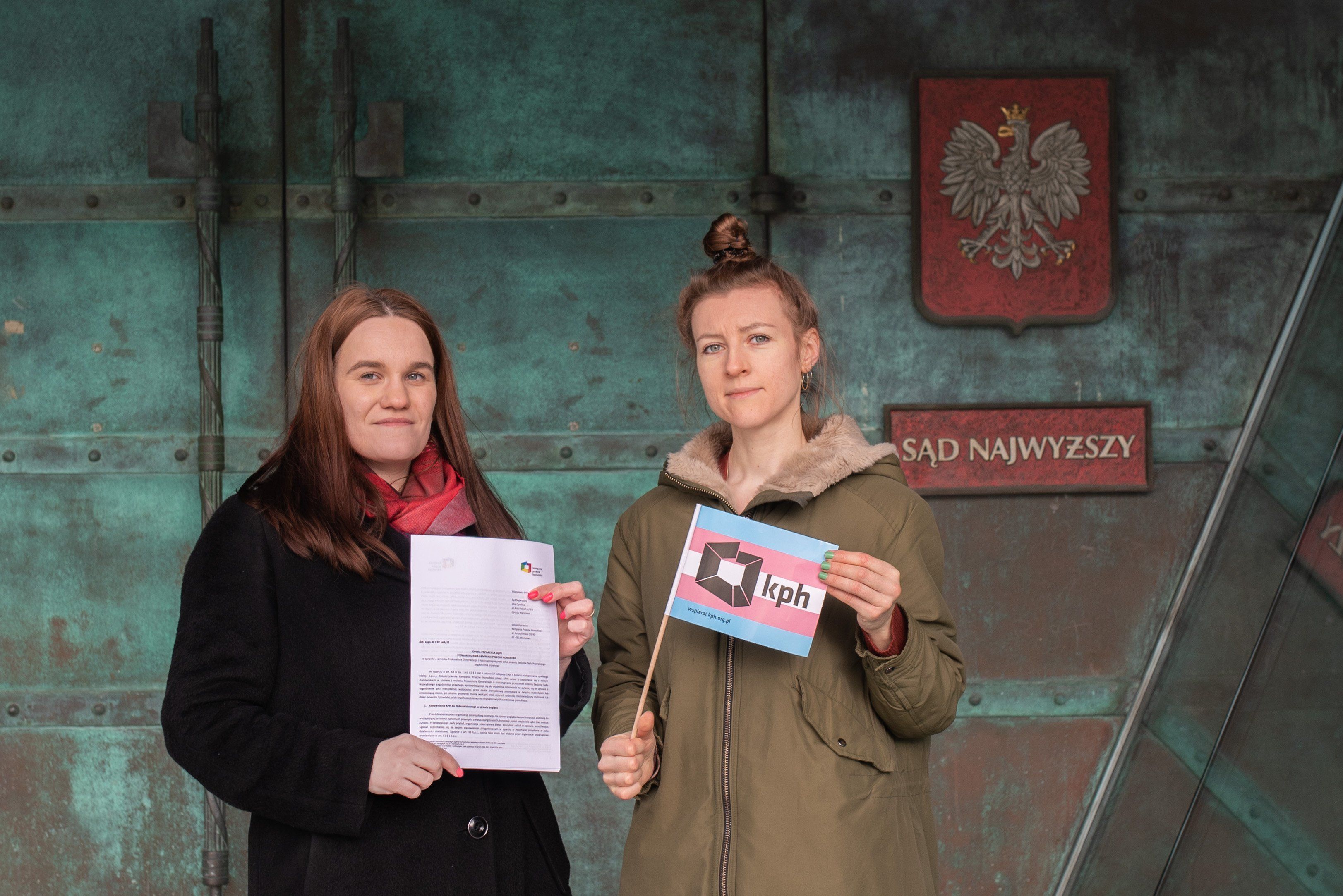 Na zdjęciu: Dwie osoby stojące przed budynkiem Sądu Najwyższego. W dłoniach trzymają opinię przyjaciela sądu i flagę w barwach osób transpłciowych