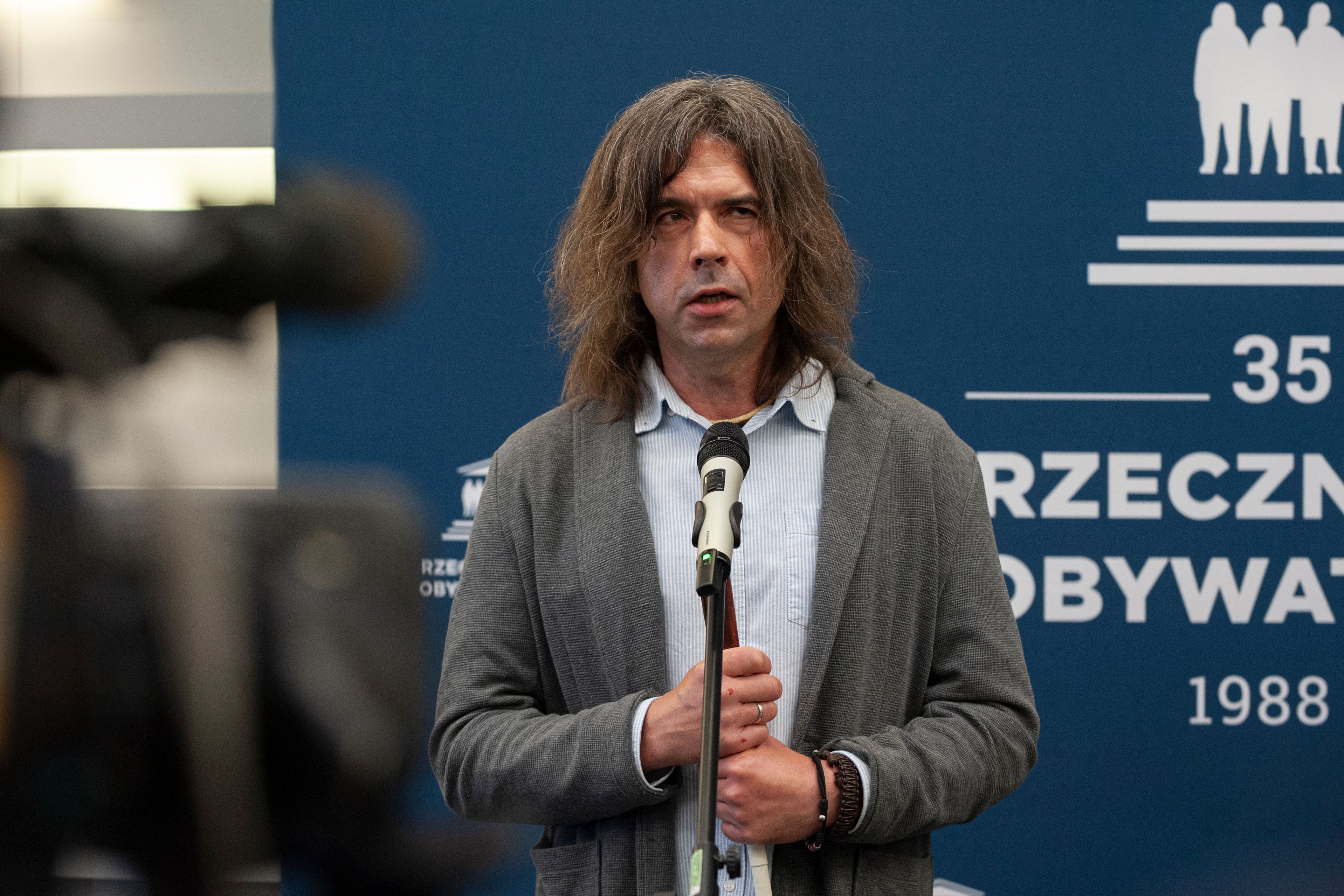 Męzczyzna w marynarce i z białą laską mówi do mikrofonu