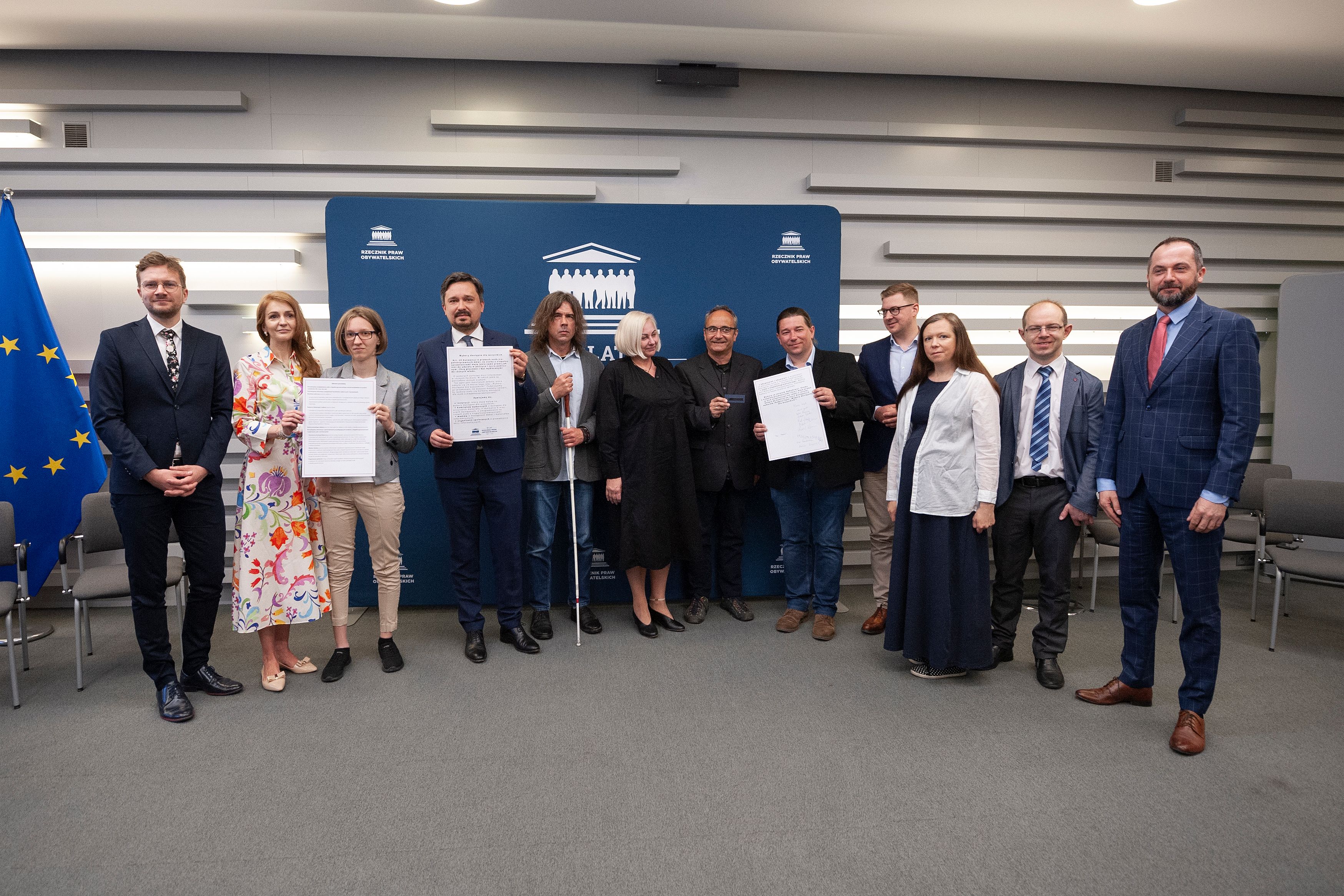 Grupa osób ( z RPO i mężczyzną z białą laską) pozuje dpo zdjęcia trzymając w rkękach kopię apelu (jest on wydtrukowany oraz oznaczony alfabetem Braille'a)