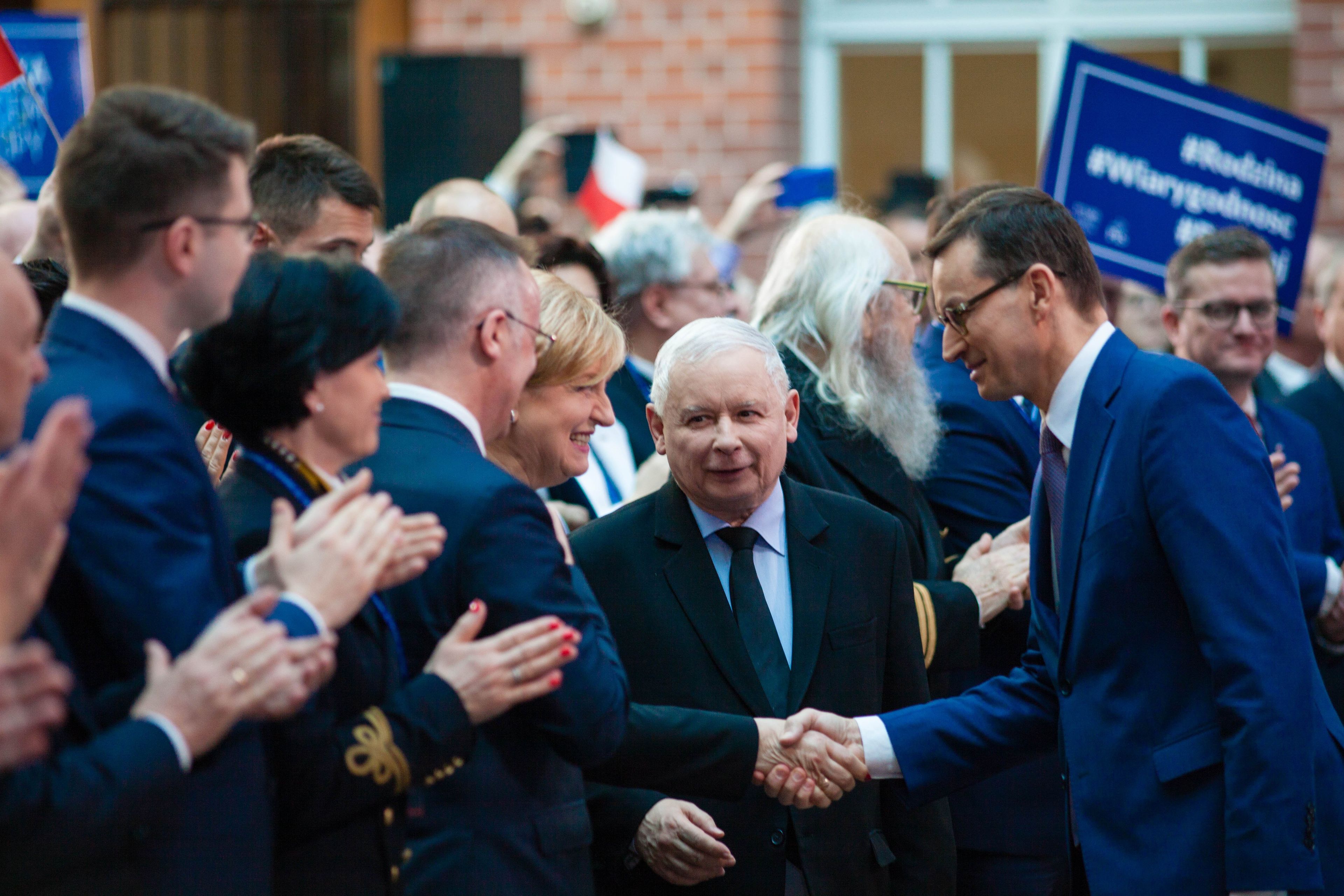 Wyższy mężczyzna z prawej ściska rękę niższego, starszego mężczyzny z lewej. Obaj są ubrani w garnitury, obok nich uśmiechnięty, wiwatujący tłum