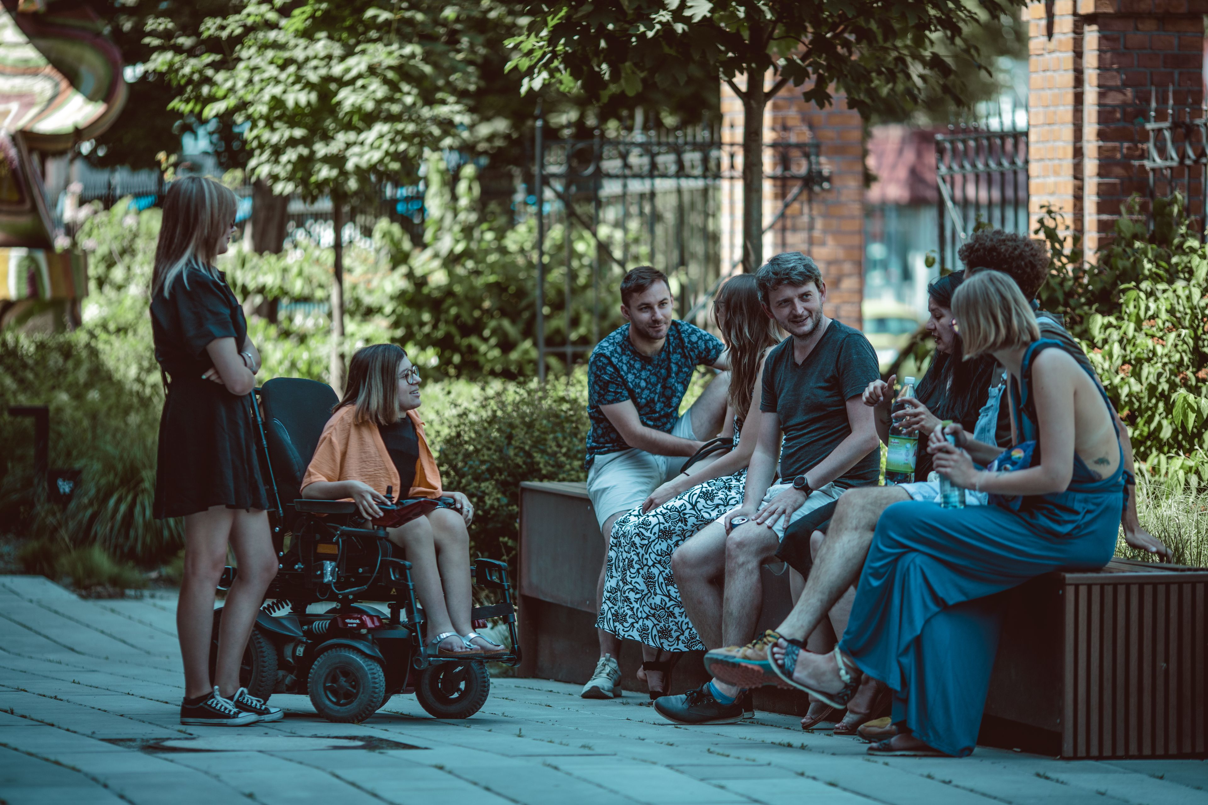 Ludzie siedzą na ławce przed budynkiem. W centrum - młody mężczyzna w ciemnej koszulce (jest lato)