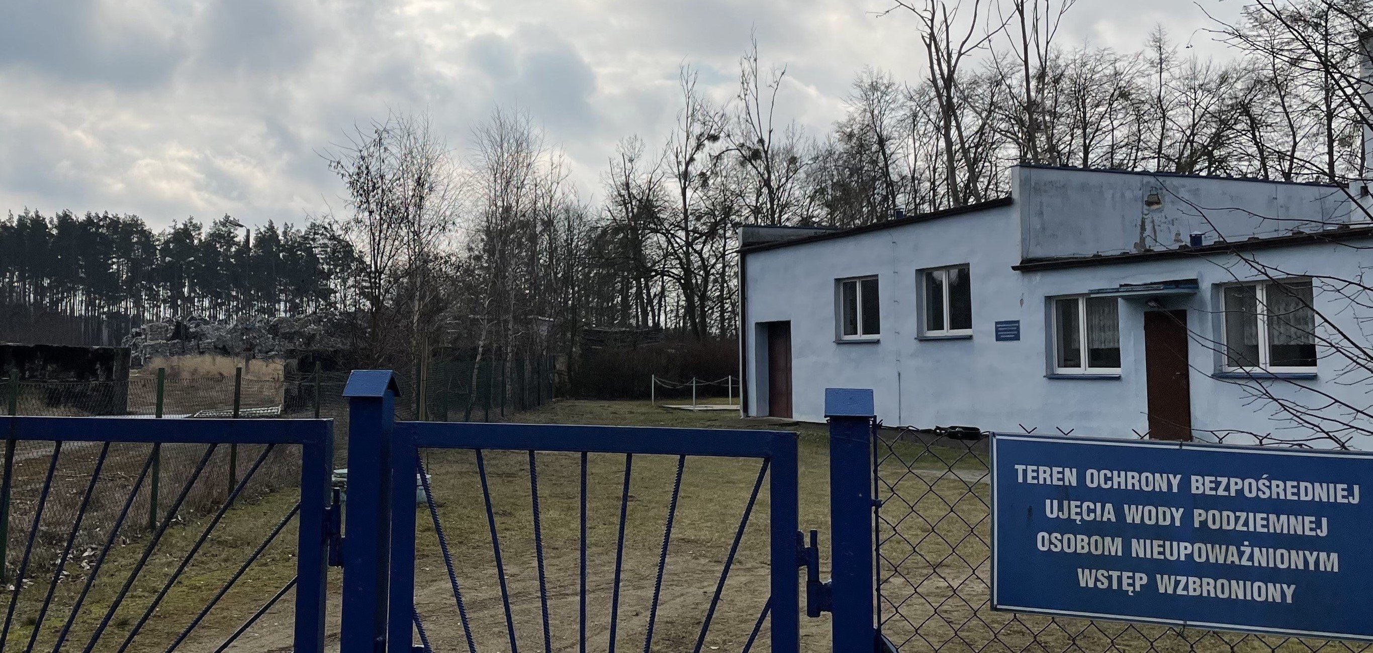 płot z niebieską tablicą: Teren ochrony bezpośredniej, ujęcie wody podziemnej, osobom nieupoważnionym wstęp wzbroniony. W głębi, za betonowym murem widoczna hałda śmieci