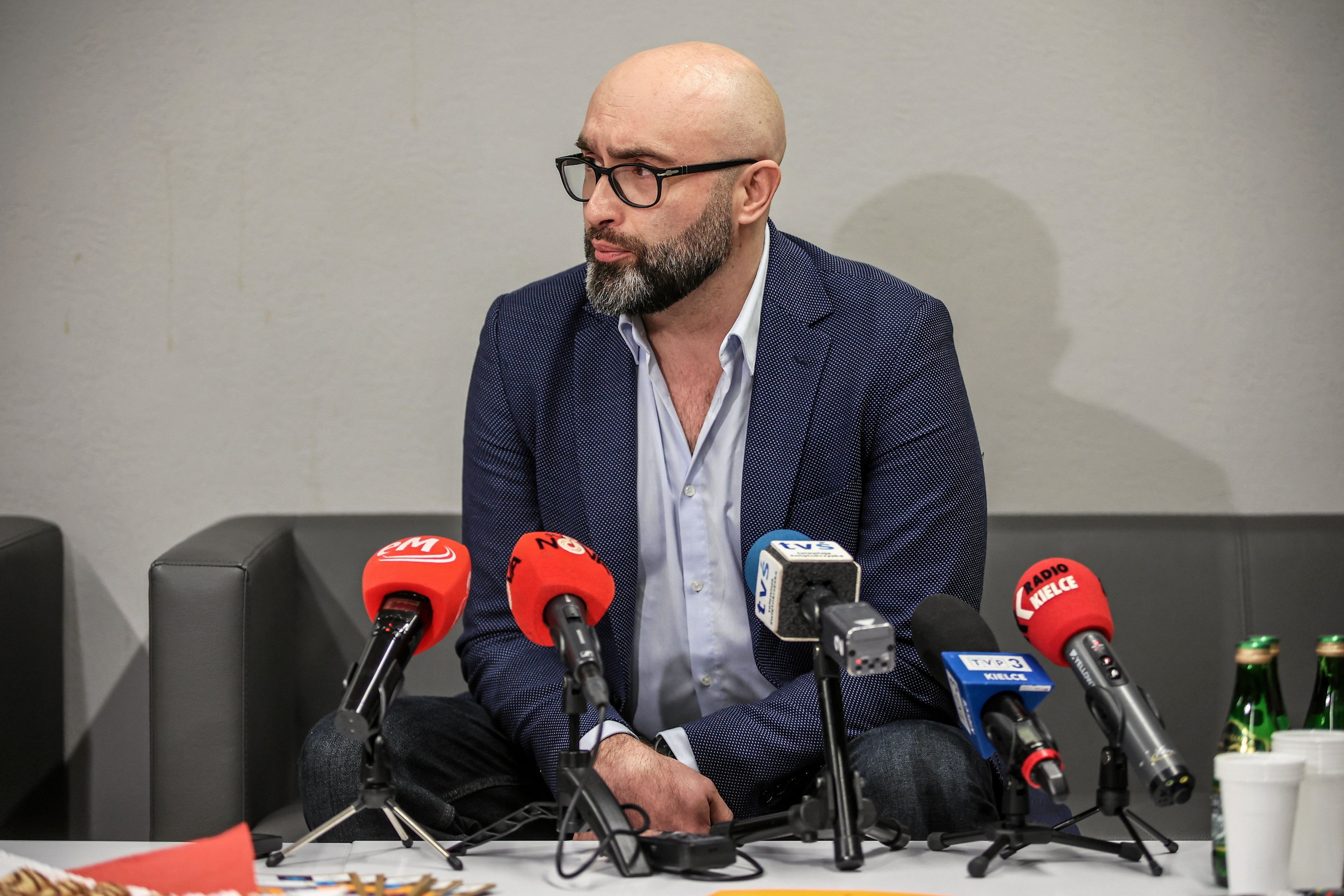 Mężczyzna z brodą w okulrach, przed nim szereg mikrofonów różnych stacji