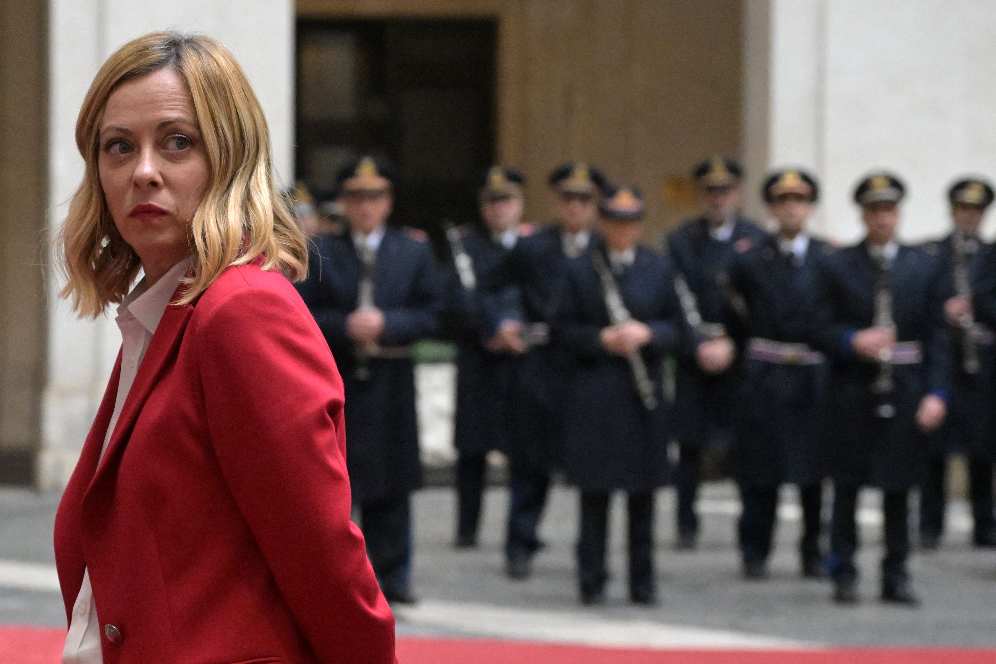 40-letnia blondynka z włosami do ramion, w czerwonym żakiecie. Za nią stoi orkiestra wojskowa. Giorgia Meloni